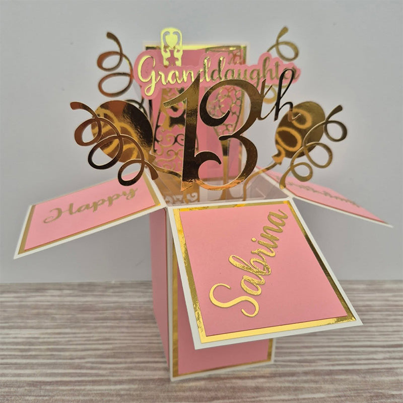 A pink and gold pop-up box birthday card with balloons and decorations springing from the box